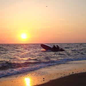 Puede incluir: Una peque&ntilde;a lancha motora inflable con dos ocupantes navegando sobre olas agitadas durante una vibrante puesta de sol. El sol se encuentra bajo en el horizonte, proyectando un c&aacute;lido resplandor dorado sobre el agua y la orilla arenosa.