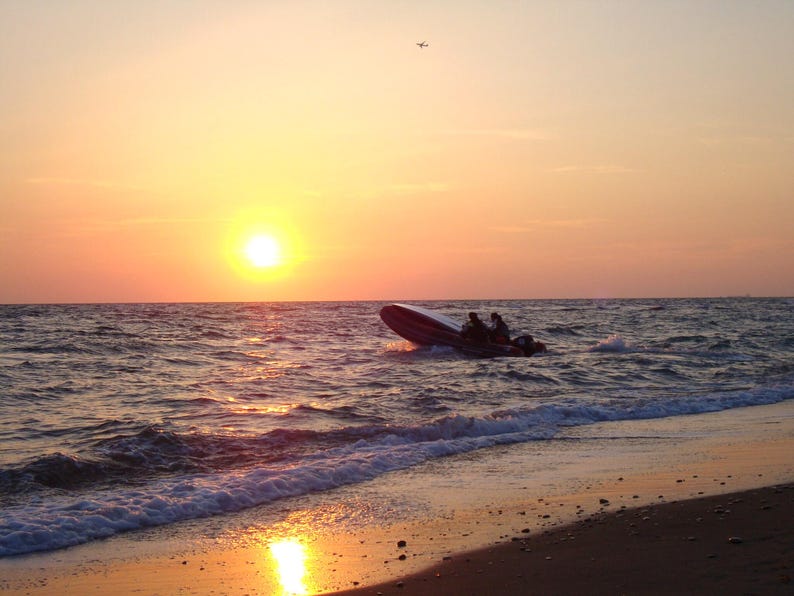 Puede incluir: Una peque&ntilde;a lancha motora inflable con dos ocupantes navegando sobre olas agitadas durante una vibrante puesta de sol. El sol se encuentra bajo en el horizonte, proyectando un c&aacute;lido resplandor dorado sobre el agua y la orilla arenosa.
