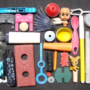 Può includere: Una collezione di giocattoli e oggetti di plastica colorati, tra cui un tubo di schiuma rosso, una molletta blu, un ponte rosa, una palla bianca, un'ala blu, una testa di bambola, una ciotola gialla, un cucchiaio rosa e una statuina di canguro.