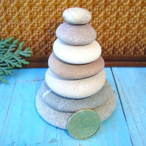 Può includere: Una pila di pietre lisce e arrotondate in varie tonalità di grigio, bianco e marrone, disposte a forma di piramide. Una moneta da due euro poggia su una superficie blu in primo piano. Lo sfondo presenta un motivo strutturato marrone dorato.