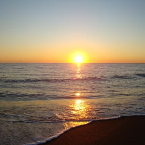 Op de afbeelding: Een levendige zonsondergang boven de oceaanhorizon waarbij de heldergele zon een gouden reflectie werpt over het kabbelende wateroppervlak. De donkere zandkust is op de voorgrond zichtbaar met wit zeeschuim van de golven.