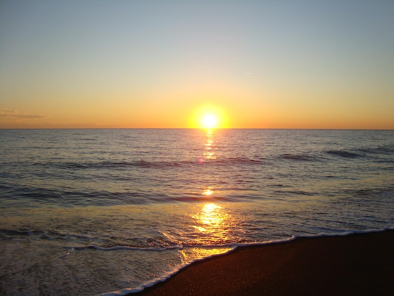 Op de afbeelding: Een levendige zonsondergang boven de oceaanhorizon waarbij de heldergele zon een gouden reflectie werpt over het kabbelende wateroppervlak. De donkere zandkust is op de voorgrond zichtbaar met wit zeeschuim van de golven.