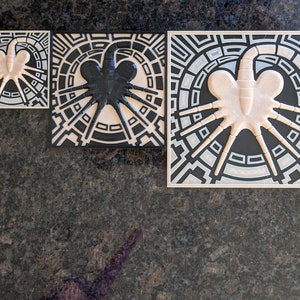 May include: Three ceramic tiles with a black and white geometric design featuring a stylized alien creature. The tiles are square and have a raised design.