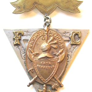 May include: Vintage Knights of Pythias medal with a triangular silver base and a central oval emblem. The emblem features a knight's helmet, crossed axes, and the inscription "ESTO PERPETUA". A gold ribbon tops the medal.