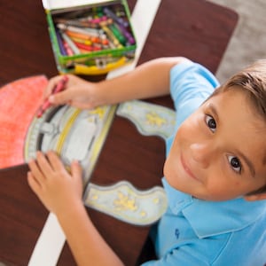 May include: A young person colors a paper craft of a helmet with a red, yellow, and blue crayon. The helmet is on a brown wooden table.