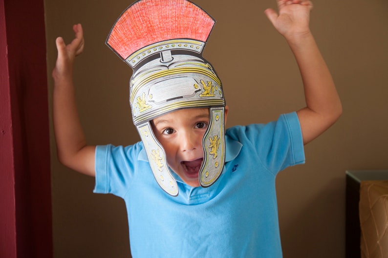 May include: A young person wearing a blue shirt and a paper Roman helmet with red and yellow accents. The helmet has a white face mask with a gold trim.