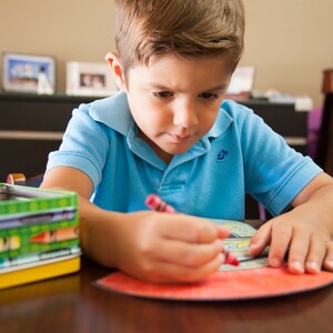 May include: A young boy sits at a table, coloring with a red crayon on a coloring page. He is wearing a blue polo shirt. A box of crayons is on the table to the left of the page.