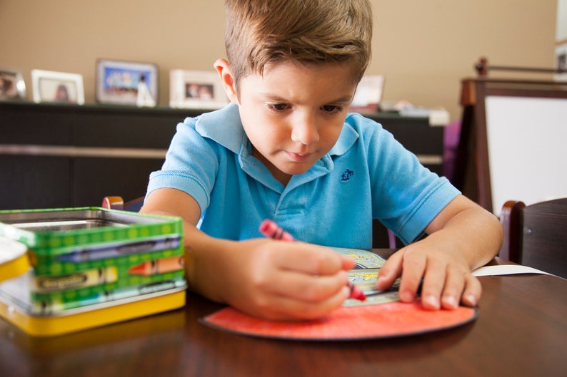 May include: A young boy sits at a table, coloring with a red crayon on a coloring page. He is wearing a blue polo shirt. A box of crayons is on the table to the left of the page.