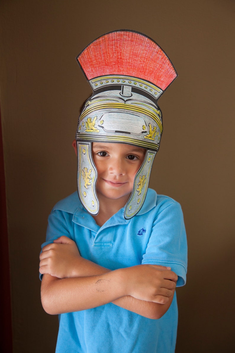 May include: A young person wearing a blue polo shirt and a paper Roman helmet with a red crest. The helmet is decorated with gold trim and a lion design.