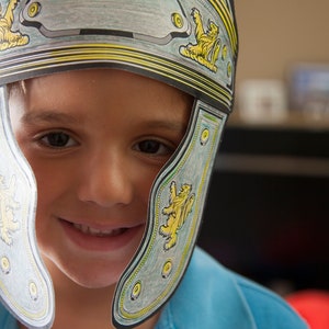 May include: A young person wearing a blue shirt and a paper Roman helmet with yellow and gold accents. The helmet has a lion design on each side.