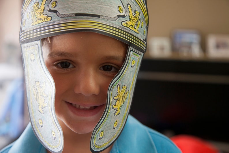 May include: A young person wearing a blue shirt and a paper Roman helmet with yellow and gold accents. The helmet has a lion design on each side.