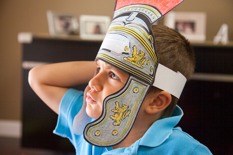 May include: A young person wearing a paper Roman helmet with a white headband. The helmet is decorated with a silver and gold design and has a red crest. The person is wearing a blue shirt.
