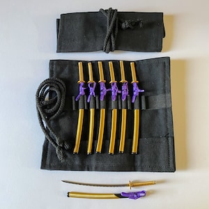 May include: A black fabric roll-up case holds six miniature gold and purple katanas. A seventh katana is unsheathed, revealing a silver blade. The case is tied with a black cord. The katanas have gold handles and purple accents.