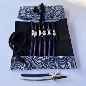 May include: A set of miniature katana swords with purple handles and silver blades, displayed in a black fabric roll with a navy and white wave pattern. The set includes a matching scabbard and a rolled-up storage case.