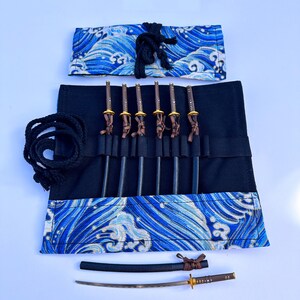 May include: A set of miniature katana swords with brown handles and gold accents, displayed in a black roll-up case with a blue wave pattern. The set includes a black cord and a matching sheath.