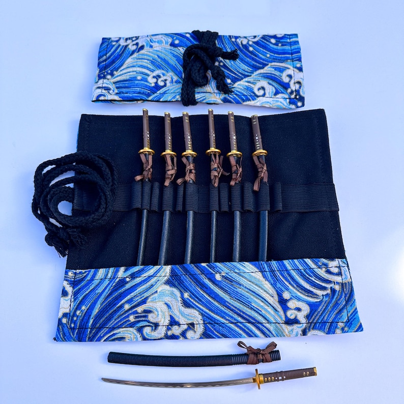 May include: A set of miniature katana swords with brown handles and gold accents, displayed in a black roll-up case with a blue wave pattern. The set includes a black cord and a matching sheath.