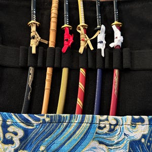 May include: A black carrying case holds six miniature katana swords. The swords have black, gold, and red accents. The case is lined with a blue and white wave pattern.