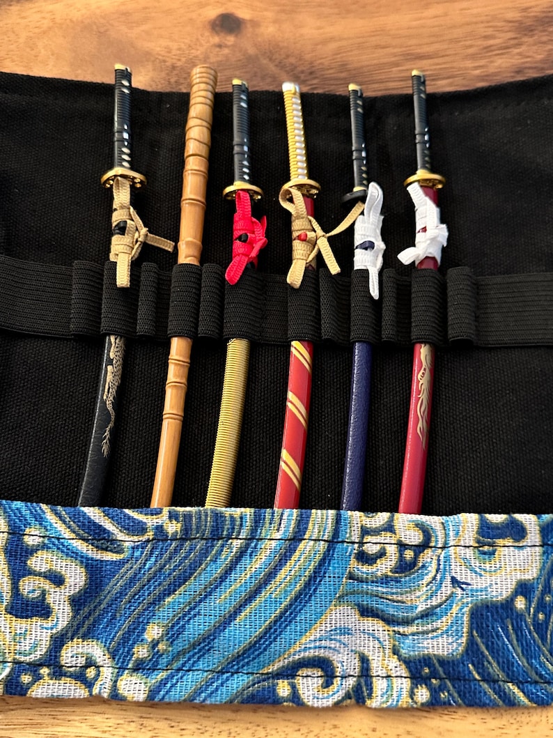 May include: A black carrying case holds six miniature katana swords. The swords have black, gold, and red accents. The case is lined with a blue and white wave pattern.