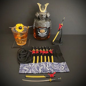 May include: A still-life arrangement featuring a bottle of Nikka Whisky in a samurai helmet-shaped holder, a cocktail, and several miniature katana swords. The cocktail is garnished with a pineapple slice. The scene has a dark background.