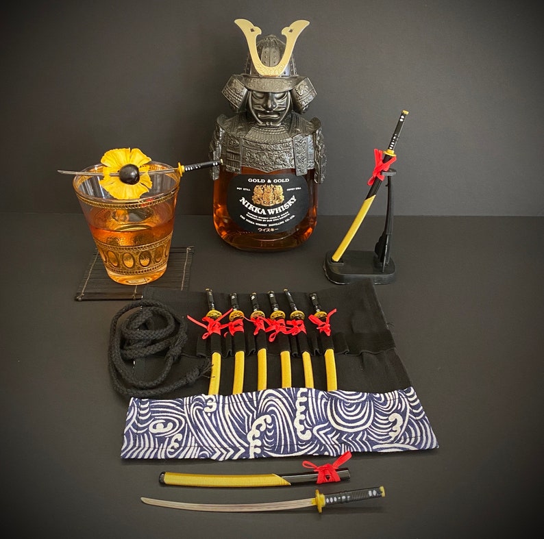 May include: A still-life arrangement featuring a bottle of Nikka Whisky in a samurai helmet-shaped holder, a cocktail, and several miniature katana swords. The cocktail is garnished with a pineapple slice. The scene has a dark background.