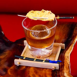 May include: A cocktail in a clear glass with a large ice sphere, garnished with a pineapple slice on a miniature sword. The drink sits on a bamboo tray, with a decorative black and gold chopstick and a blue ribbon. The background is a solid red.