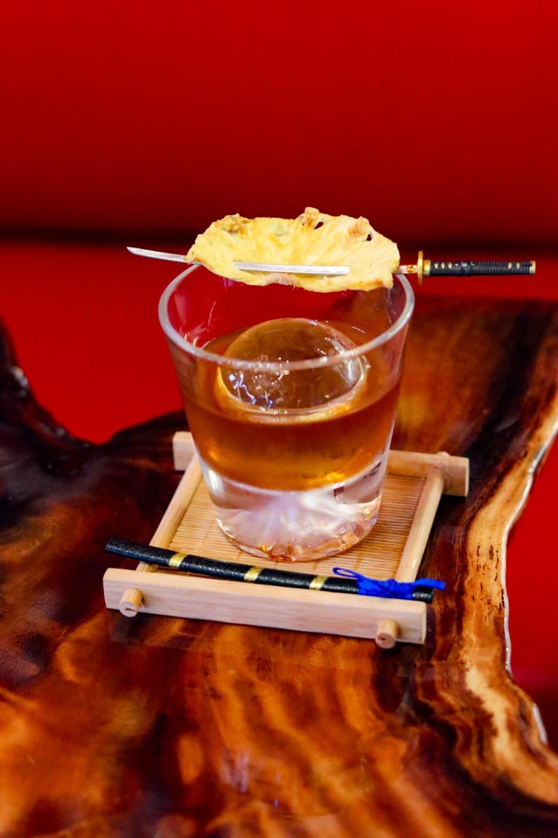May include: A cocktail in a clear glass with a large ice sphere, garnished with a pineapple slice on a miniature sword. The drink sits on a bamboo tray, with a decorative black and gold chopstick and a blue ribbon. The background is a solid red.