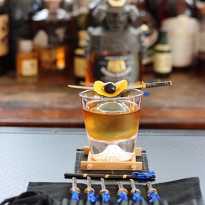May include: A cocktail in a rocks glass with a large ice sphere, garnished with a cherry and orange peel. The glass sits on a bamboo coaster with a mountain design. A set of miniature katana-style cocktail picks are displayed.