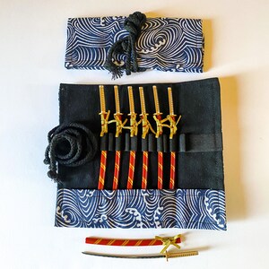 May include: A black fabric roll with a blue and white wave pattern, holding miniature gold and black swords and red and gold sticks. A black rope and a matching fabric wrap are also included. A miniature sword and ruler are at the bottom.