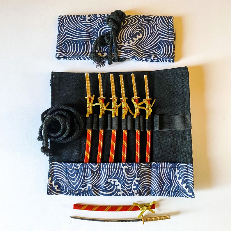 May include: A black fabric roll with a blue and white wave pattern, holding miniature gold and black swords and red and gold sticks. A black rope and a matching fabric wrap are also included. A miniature sword and ruler are at the bottom.