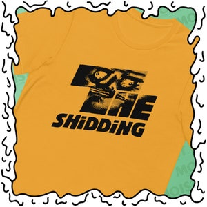 May include: Mustard yellow t-shirt with a black graphic design featuring a distorted face and the text "THE SHIDDING". The shirt is made of a soft material and has a crew neck.
