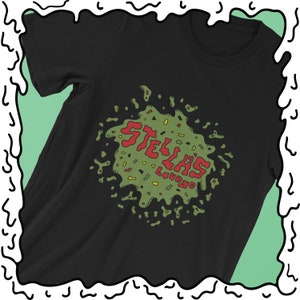 May include: Black t-shirt featuring a colorful graphic design. The design includes the text "STELLA'S Lounge" in red, with a green and yellow splatter-like background. The shirt has a crew neck and short sleeves.
