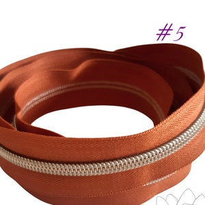 May include: A close-up of a rust-coloured zipper with a silver zipper pull. The zipper is folded in a circle and has a white stripe running along the edge.