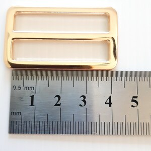 K&ouml;nnte beinhalten: Goldfarbene rechteckige Schiebeschnalle mit zwei &Ouml;ffnungen. Die Schnalle befindet sich neben einem Lineal, das Messungen in Millimetern anzeigt. Die Schnalle ist etwa 5 cm lang.