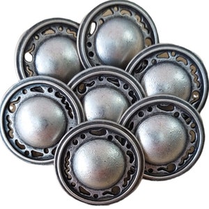 May include: A group of seven silver-toned metal buttons with a decorative, ornate design around the edge. The buttons are round and have a raised centre.