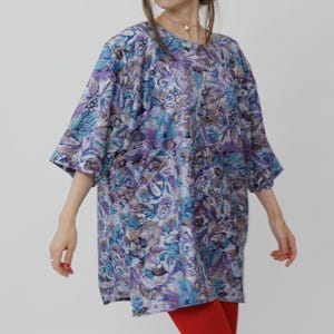 Op de afbeelding: Een ruimvallende tuniek met een levendig bloemenpatroon in tinten paars, blauw en groenblauw, met ellebooglange mouwen en een ronde halslijn, gecombineerd met een effen felrode ondoorzichtige panty die de benen en voeten volledig bedekt.