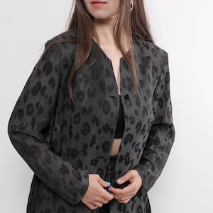 May include: A dark gray blazer with a subtle leopard print pattern. The long-sleeved jacket has a collarless design and a button closure. The model is wearing black jeans and a black top. The blazer is a stylish addition to any wardrobe.