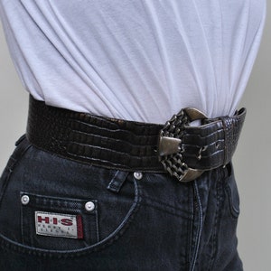 May include: A pair of dark wash denim jeans with a brown crocodile embossed belt and a silver buckle. The belt buckle has a unique design. The jeans have a small red and white patch on the front pocket that says "HIS Henry I. Siegel".