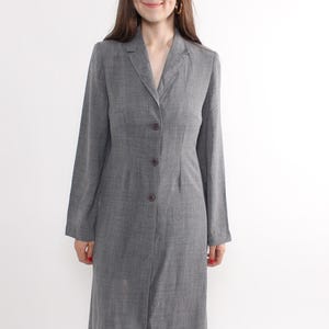 May include: A long, gray blazer-style dress with a button-down front. The dress has long sleeves and a tailored fit. The person is wearing black shoes. The background is plain white.