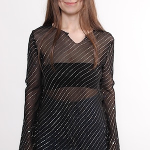 Può includere: Top nero trasparente con strisce diagonali argentate e maniche lunghe. Il top ha uno scollo a V e una vestibilità ampia e fluida. La modella indossa jeans blu.