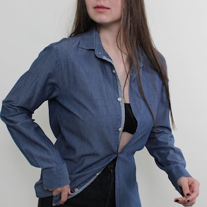 May include: A denim blue button-down shirt with long sleeves. The shirt is unbuttoned, revealing a black bra. The shirt has a classic collar and cuffs. The model is wearing black pants.