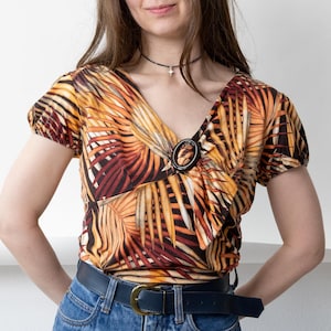 Könnte beinhalten: Kurzärmliges Oberteil mit V-Ausschnitt und tropischem Blattmuster in Orange-, Gelb- und Brauntönen. Dazu eine blaue Jeans und ein dunkelblauer Gürtel mit goldener Schnalle. Ein schwarzes Choker-Halsband rundet das Outfit ab.