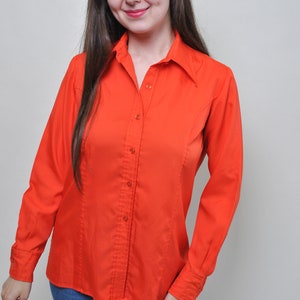 May include: Bright orange button-down shirt with long sleeves. The shirt has a classic collar and is paired with blue jeans. The shirt is made of a smooth fabric and has vertical seams for a tailored fit. A vintage style.