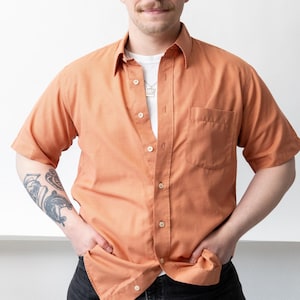 May include: An orange short-sleeved button-down shirt with a chest pocket. The shirt is worn over a white t-shirt and paired with black jeans. The person has a moustache and tattoos on their arm.
