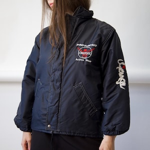 May include: A dark navy blue windbreaker jacket featuring a high collar, long sleeves with elastic cuffs, and a front zipper closure. The chest displays white and red text reading WINDSURFING CHIEMSEE Active Wear, with the word Chiemsee on the sleeve.
