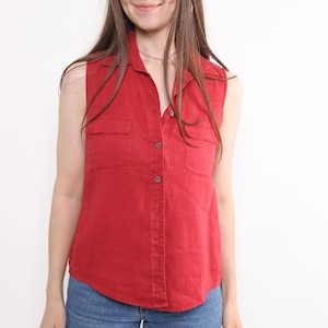 May include: A sleeveless red shirt with two chest pockets and a button-down front. The shirt is paired with blue jeans. The model is smiling, and the background is plain white.