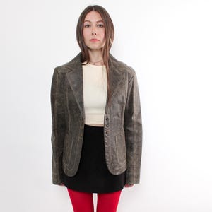 Puede incluir: Chaqueta de cuero marr&oacute;n de estilo vintage con solapas y cierre de botones. El conjunto incluye una camiseta crema, una falda negra, medias rojas brillantes y zapatillas negras. El conjunto se presenta sobre un fondo blanco.