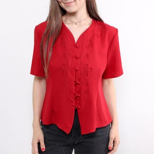 May include: A bright red short-sleeved blouse with a V-neck and embroidered detailing. The blouse has small buttons down the front and is made of a smooth material. The top is worn with black jeans.