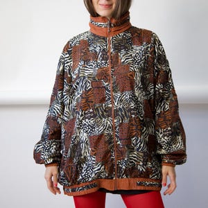 Könnte beinhalten: Eine Reißverschlussjacke mit einem gemusterten Design aus braunen, schwarzen und weißen Tierdrucken und geometrischen Formen. Die Jacke hat einen hohen Kragen, lange Ärmel mit elastischen Bündchen und einen einfarbigen braunen Saum am Reißverschluss.