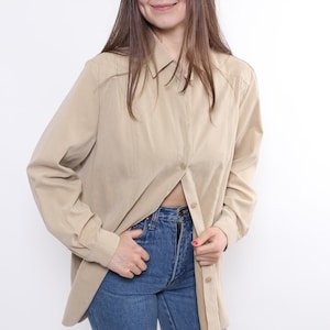 May include: A tan button-down shirt with long sleeves. The shirt is unbuttoned at the midriff, revealing blue jeans. The shirt has a classic collar and is made of a smooth fabric. The overall style is casual and relaxed.
