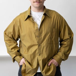 Puede incluir: Una camisa abotonada de color mostaza con mangas largas y un bolsillo en el pecho. La camisa se lleva sobre una camiseta blanca y se combina con vaqueros negros. El modelo está de pie con las manos en los bolsillos.